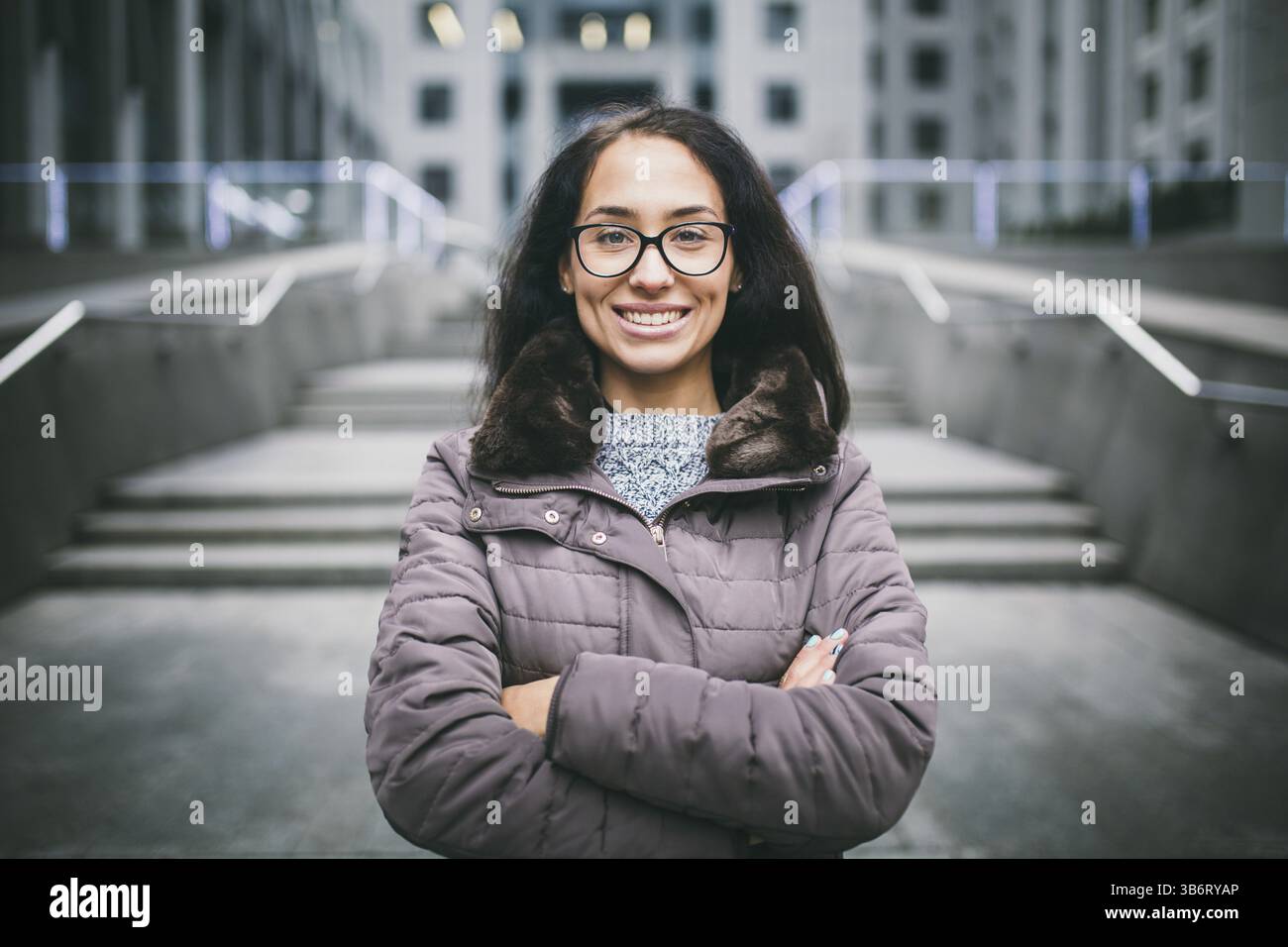 Bella giovane donna di etnia europea con bruna lungo i capelli con un sorriso toothy, indossare occhiali e un attaccapanni contro il fondale di una busin Foto Stock