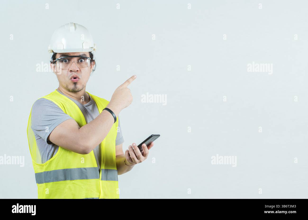 Sorpreso il lavoratore del costruttore con il telefono che puntava a uno spazio vuoto isolato. Stupito ingegnere che tiene il telefono e punta il lato isolato Foto Stock