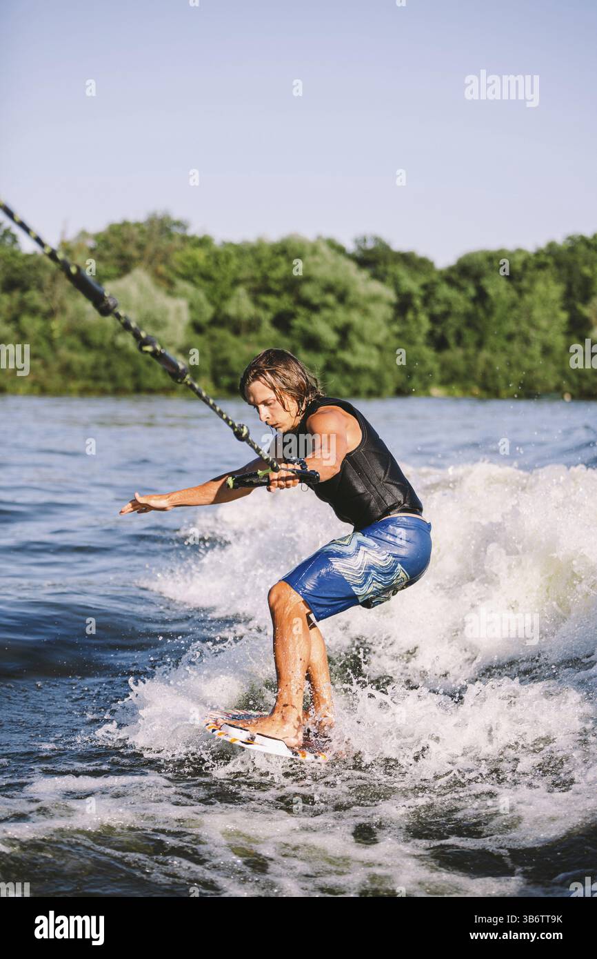 Bella vesurf biondo con capelli lunghi in gilet a bordo lungo le onde del lago. Atleta atleta maschile atletica risveglia il surf in estate sul fiume. Wakesurf acqua di spruzzata Foto Stock