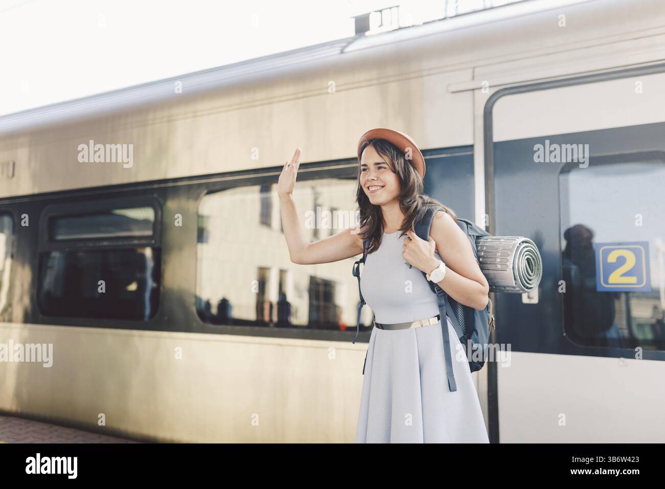 Trasporto e viaggio a tema. Ritratto di una giovane donna caucasica con sorriso in piedi alla stazione ferroviaria sfondo con zaino che saluta ciao W Foto Stock