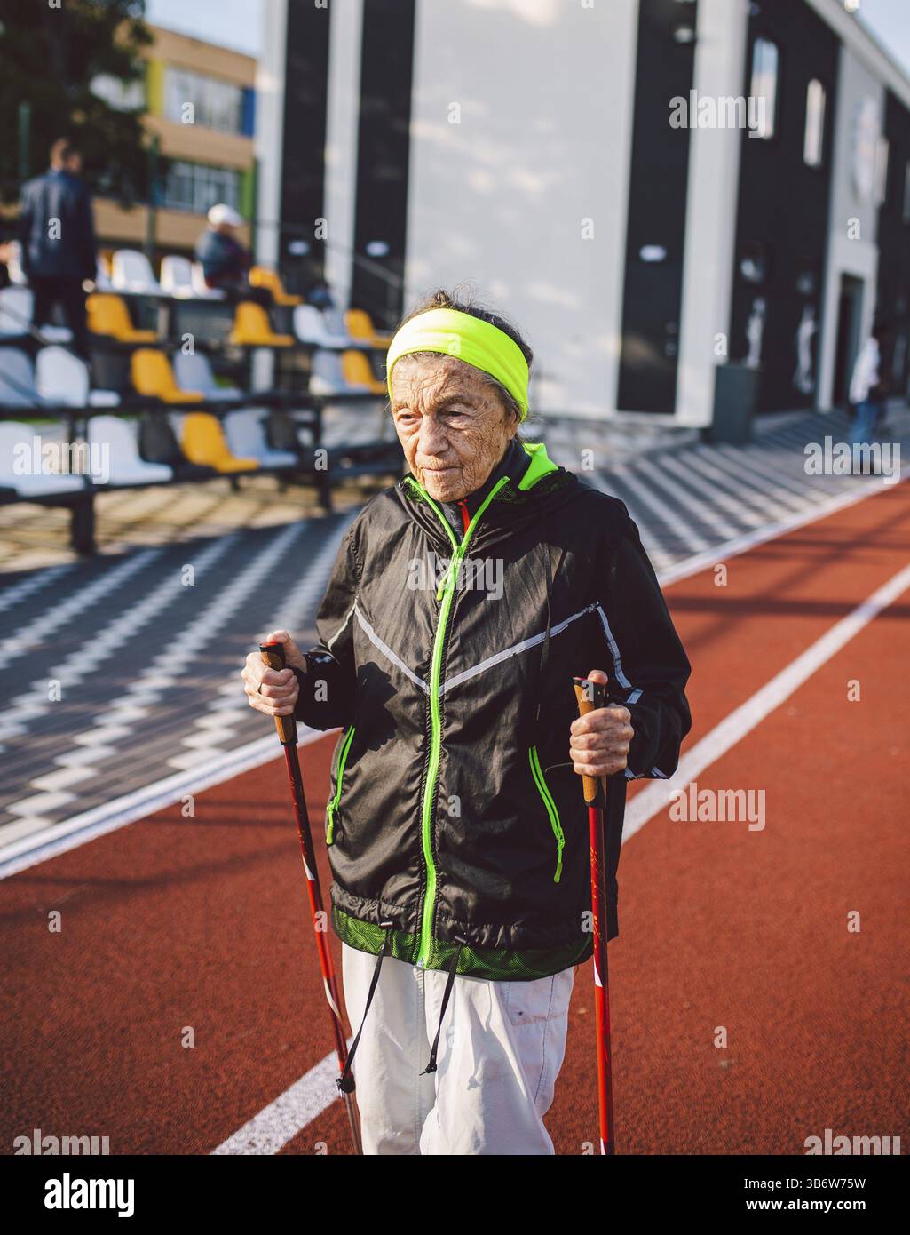 Donna anziana in abbigliamento sportivo che pratica nordic walking all'aperto su un tapis roulant in gomma nello stadio. Le donne più anziane camminano a piedi in scandinavia utilizzando il bastone da trekking Foto Stock