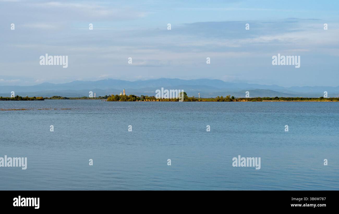 Vista distante dell'Isola di Barbana con la sua iconica Chiesa circondata da acque calme e sfondo montano a grado, Italia Foto Stock