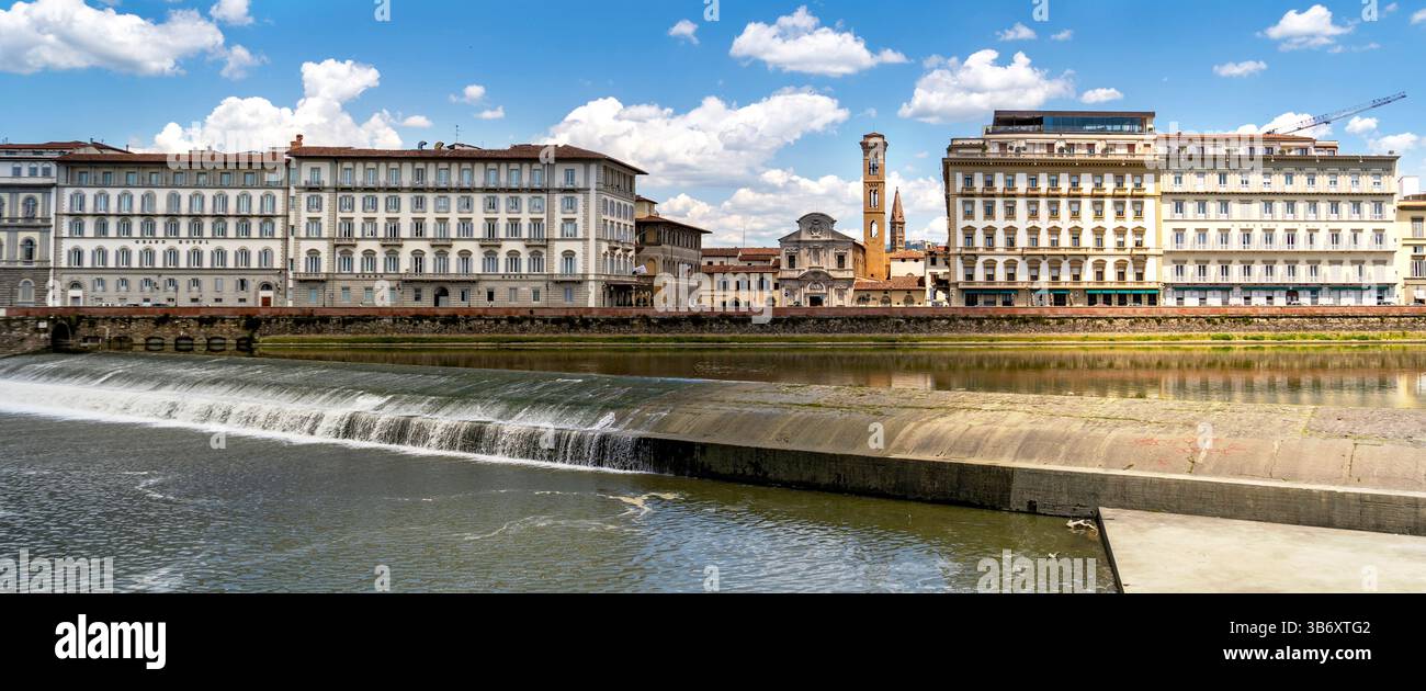 Una vista panoramica di un fiume con una cascata in primo piano, fiancheggiata da edifici storici e una torre dell'orologio sotto un cielo blu con soffici nuvole. Foto Stock