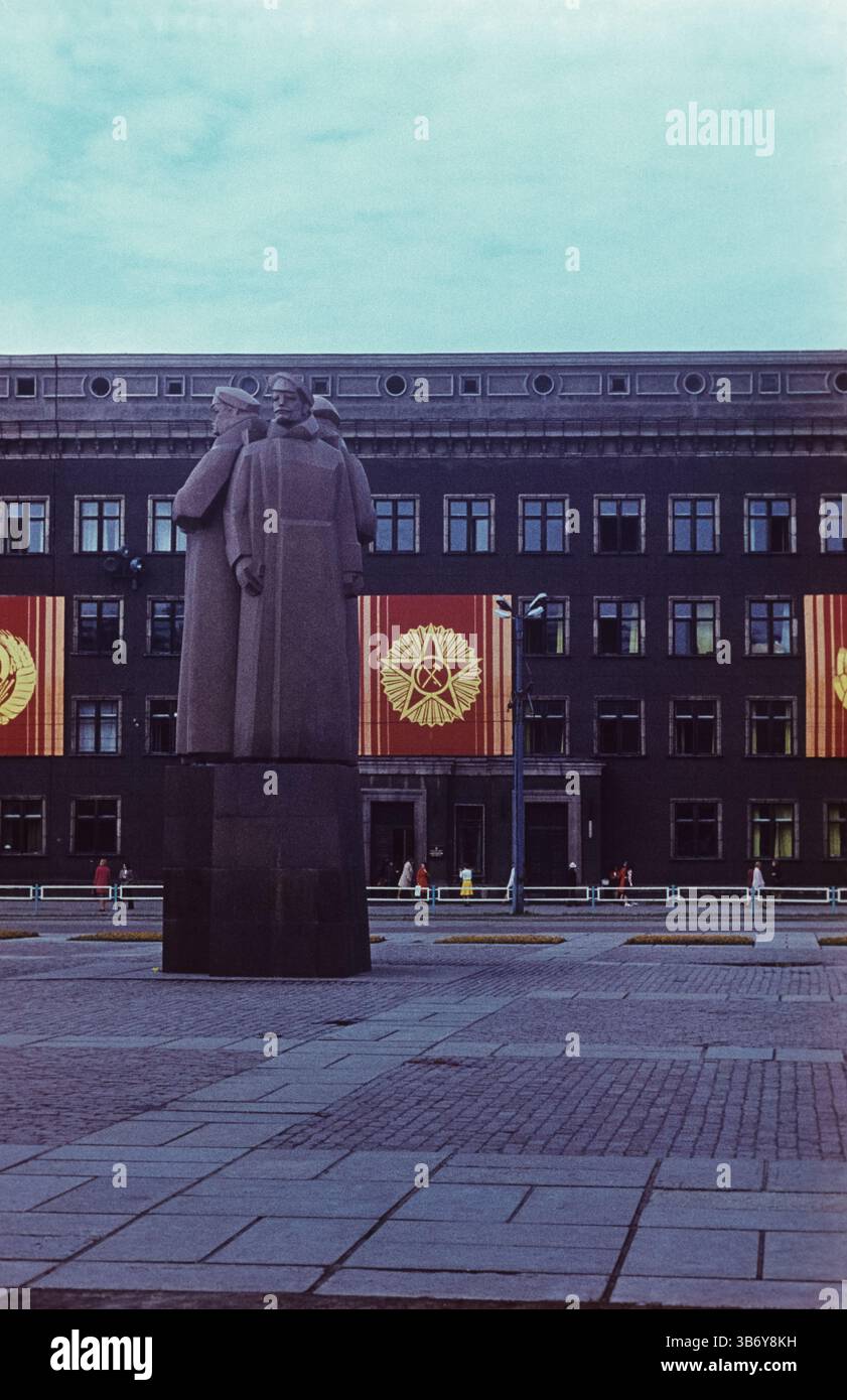 Il Monumento dei Riflemen lettoni dell'era sovietica a riga si erge davanti a un edificio governativo drappeggiato da emblemi comunisti, che riflette il passato sovietico della Lettonia dal t Foto Stock