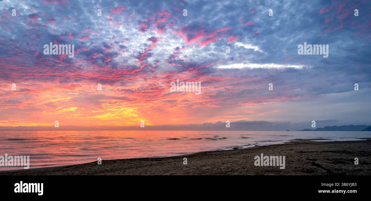 Una tranquilla scena di spiaggia al tramonto, caratterizzata da colori vivaci nel cielo con nuvole che riflettono sfumature di arancione, rosa e viola. Il mare calmo rispecchia Foto Stock