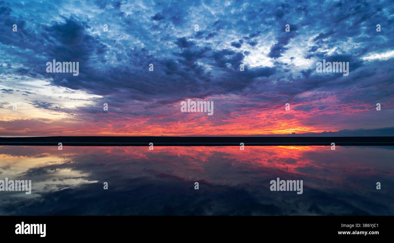 Uno splendido tramonto su un calmo corpo d'acqua, che riflette colori vivaci nel cielo. La scena presenta nuvole spettacolari e un paesaggio sereno che crea Foto Stock