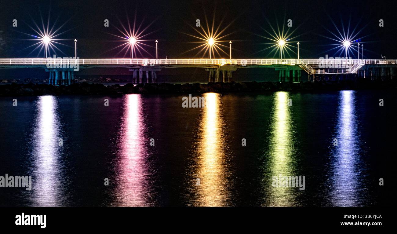 Una vista notturna di un molo illuminato da luci colorate che si riflettono sulla superficie dell'acqua. La scena cattura un'atmosfera serena con stelle vibranti Foto Stock