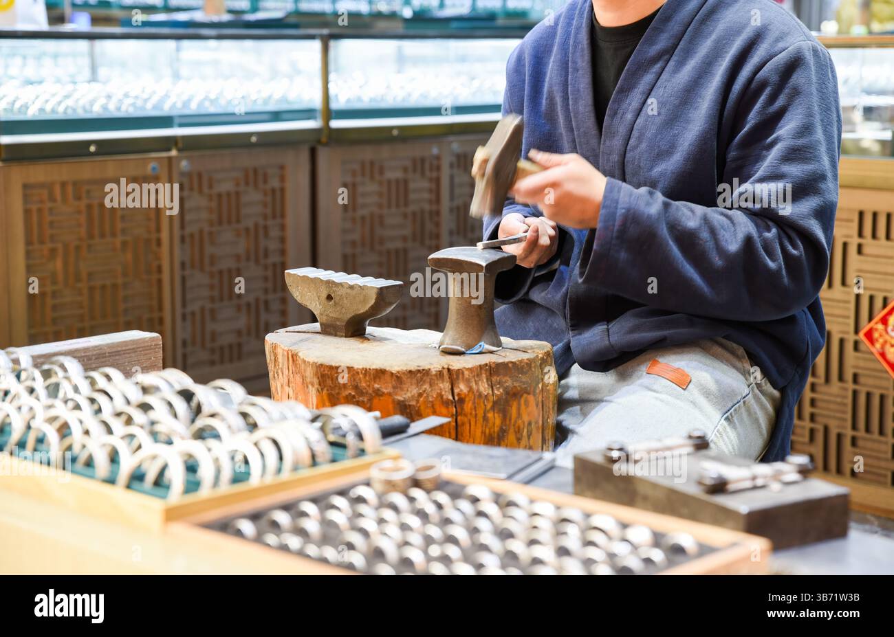 Un artigiano in una tradizionale giacca blu crea a mano anelli di gioielli su un'incudine nella Old Street Yunnan di Shanghai, con anelli in metallo finiti esposti in W Foto Stock