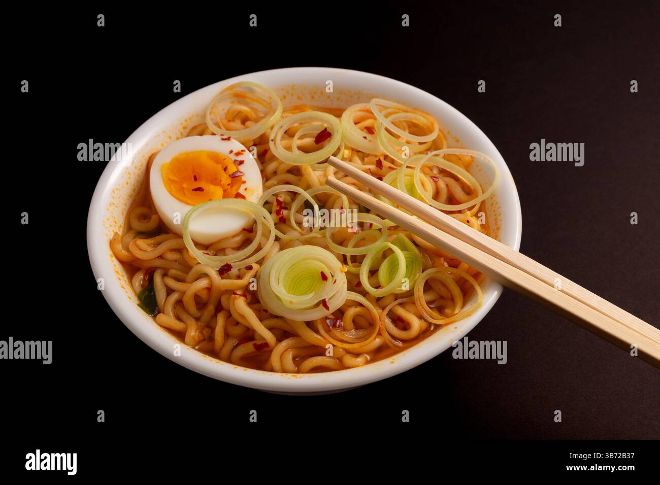 Vista dall'alto della zuppa di ramen con spaghetti che nuotano nel brodo infuso di peperoncino con fette di porro e uova, bacchette pronte per l'azione. Sfondo nero. Foto Stock