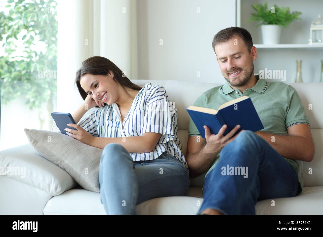 Coppia felice leggendo eBook e libro cartaceo all'interno di una casa Foto Stock