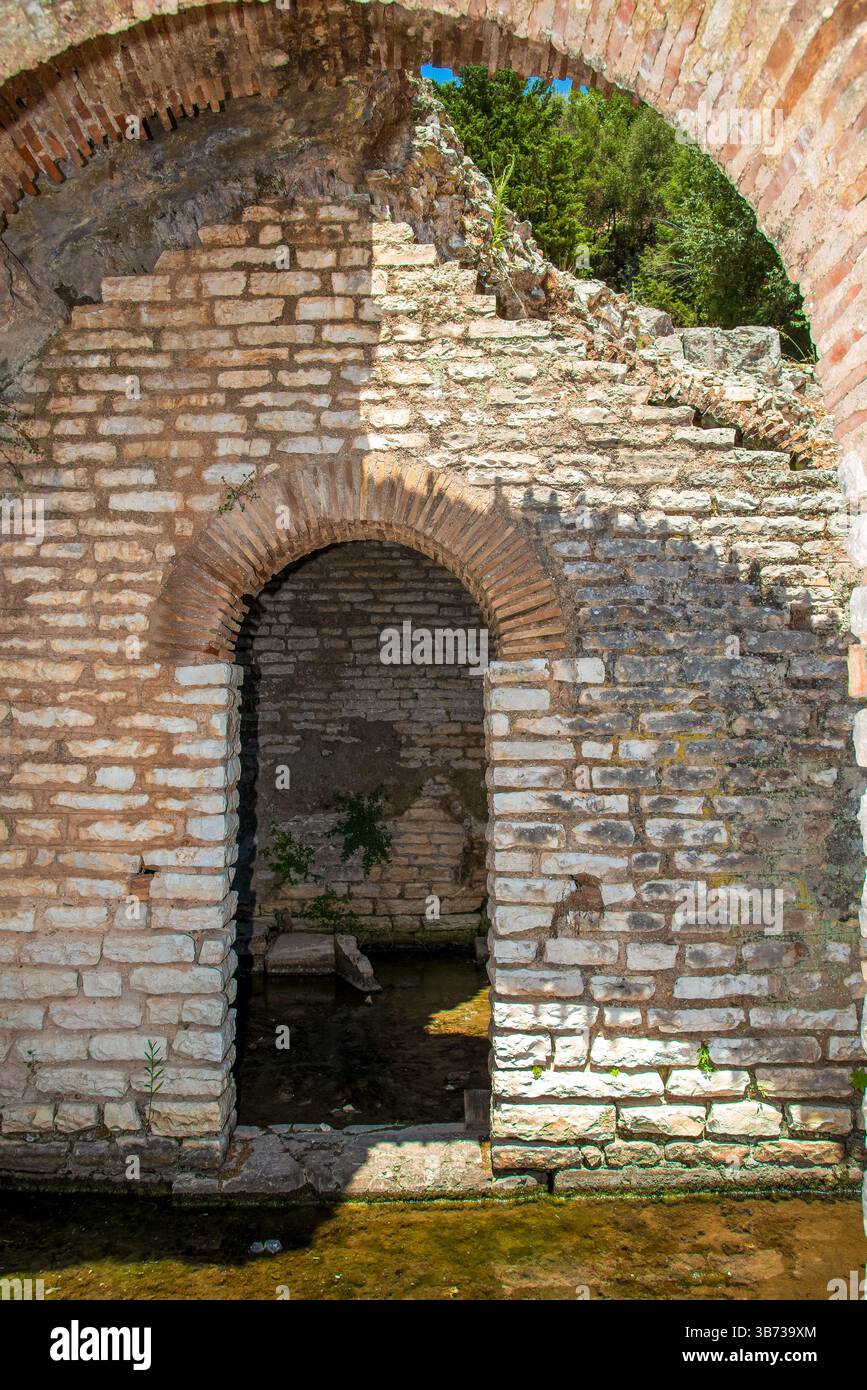 Antica arcata di pietra calcarea illuminata dal sole ai margini di un canale d'acqua riflettente all'interno delle rovine archeologiche di Butrint nel sud dell'Albania Foto Stock
