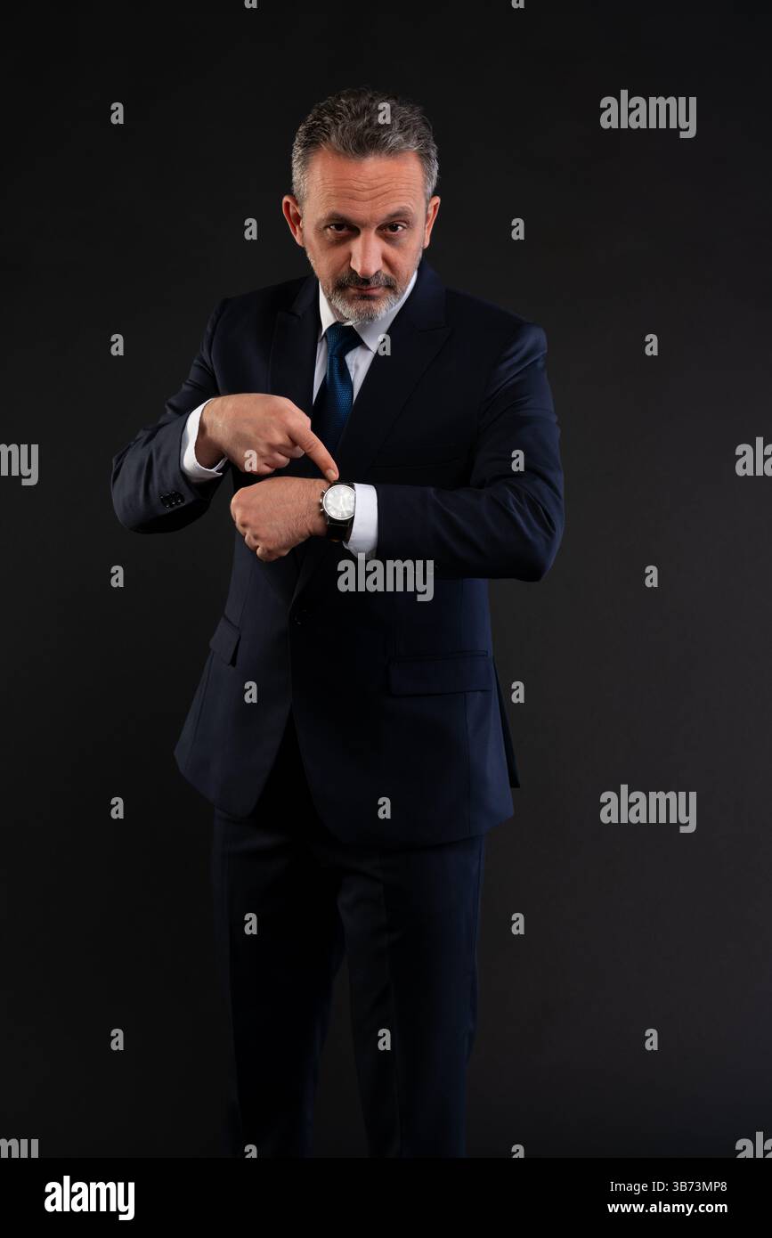 Uomo d'affari maturo in tuta che indica il suo orologio da polso, esprime urgenza o ricorda il tempo e la puntualità. Foto Stock