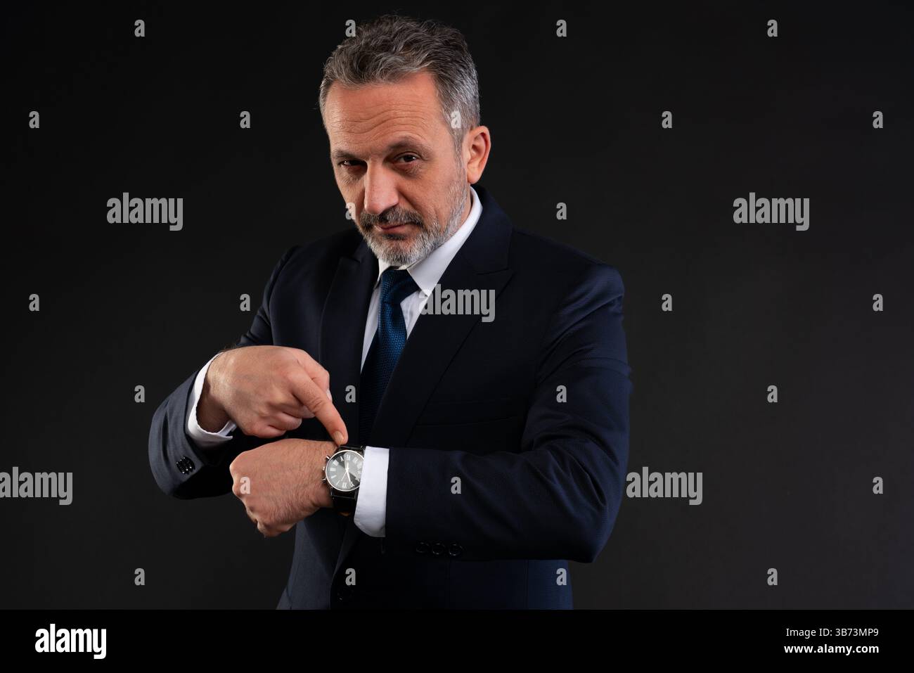 Uomo maturo in abito formale che indica il suo orologio con un'espressione sicura e assertiva, evidenziando la consapevolezza del tempo o l'urgenza. Foto Stock