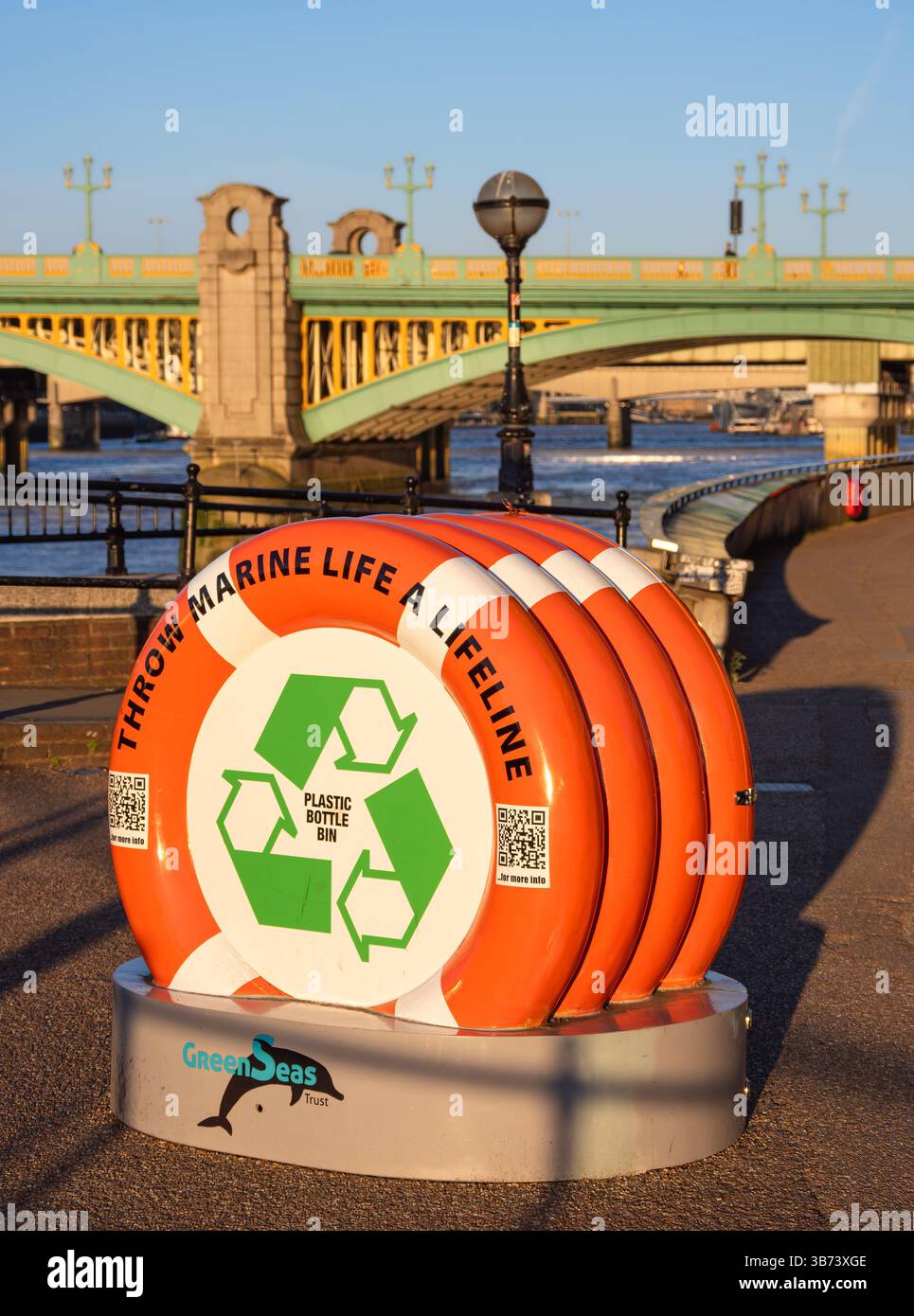 Londra, Regno Unito - 28 aprile 2025: Bidone in plastica sotto forma di una fila di salvagente arancioni con simboli di riciclaggio e un ponte sullo sfondo Foto Stock