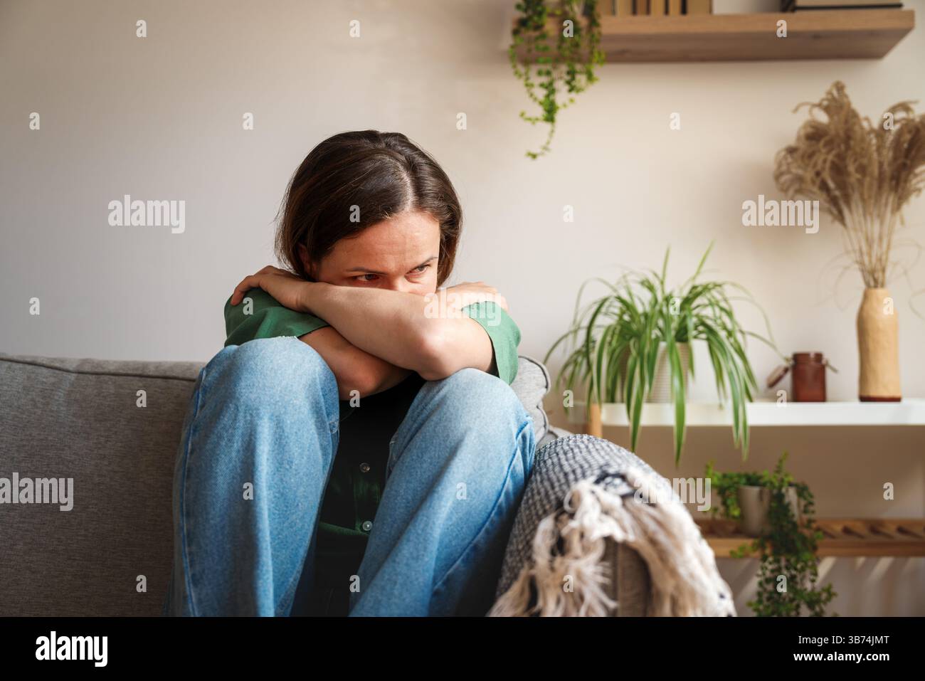 Donna triste seduta in posizione fetale mentre si sente emotivamente sopraffatta a casa Foto Stock