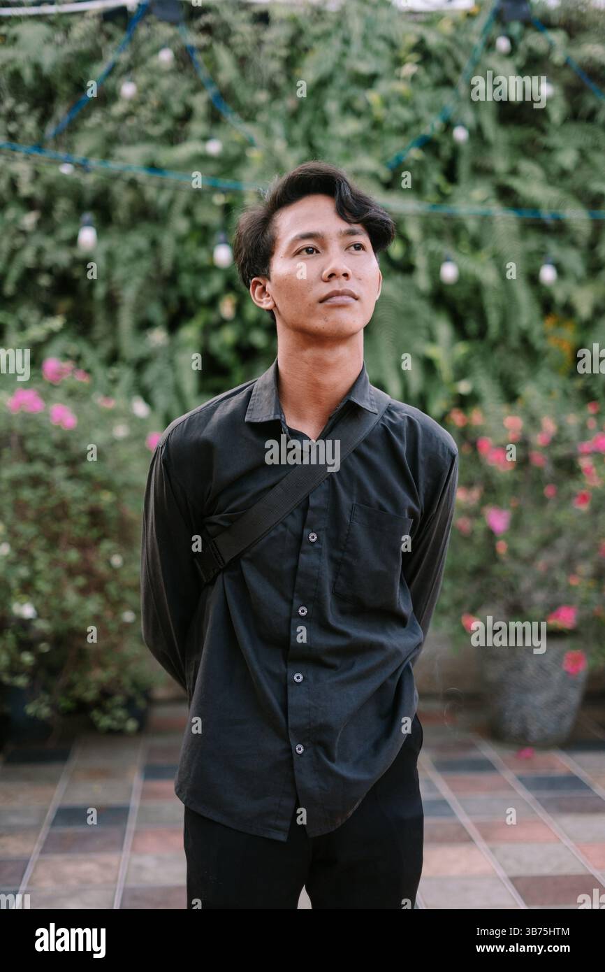 Un giovane e premuroso uomo asiatico posa all'aperto in un abbigliamento casual ed elegante, circondato da lussureggiante vegetazione e fiori. Foto Stock