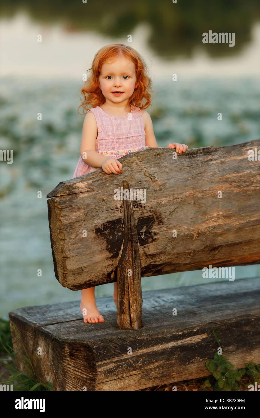 Allegra bambina dai capelli rossi con un abito rosa in piedi nudi su una grande panchina rustica in legno, tenendola giocosamente sullo schienale e sbirciando fuori wi Foto Stock