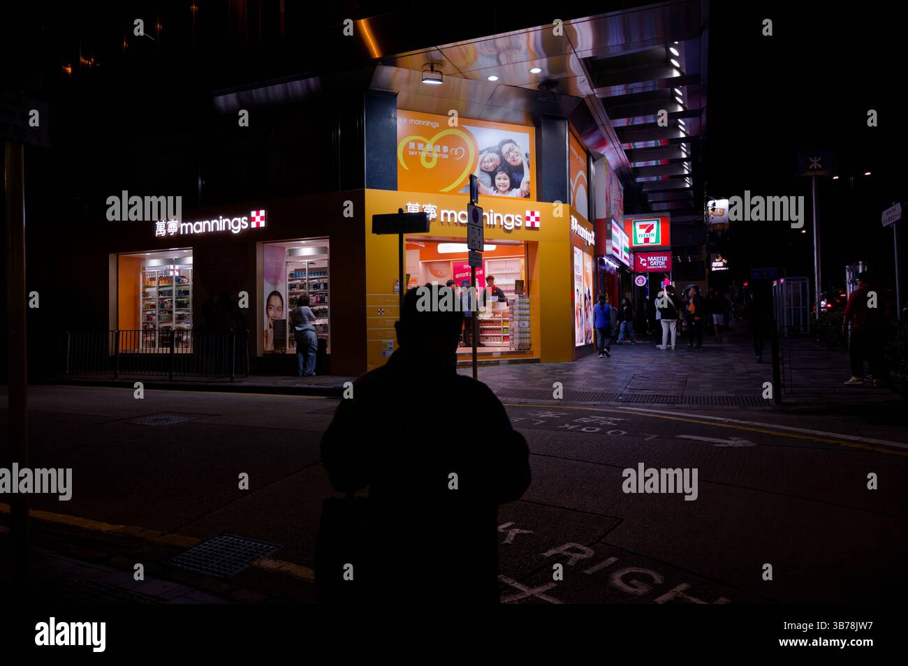 Lone silhouette si ferma dall'altra parte della strada mentre le luci luminose provenienti da una farmacia illuminano un angolo vivace Foto Stock