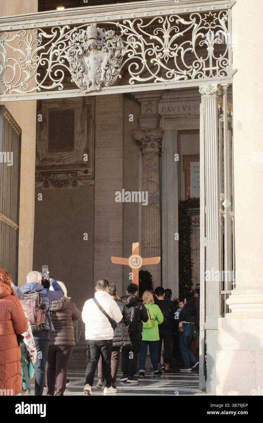 Pellegrini e turisti che entrano nella porta Santa della Basilica di San Pietro nella città del Vaticano attraverso le porte di bronzo ornate durante l'evento del Giubileo dell'anno Santo con croce visibile e raduno spirituale presso il sito sacro cattolico. Foto di alta qualità Foto Stock