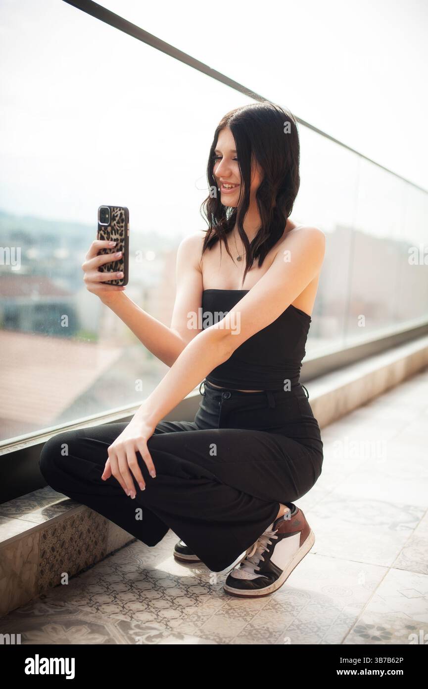 Una giovane ragazza alla moda è seduta su una sporgenza elegante e sorride mentre scatta un selfie con il telefono. Lo sfondo mostra uno splendido paesaggio urbano, Foto Stock