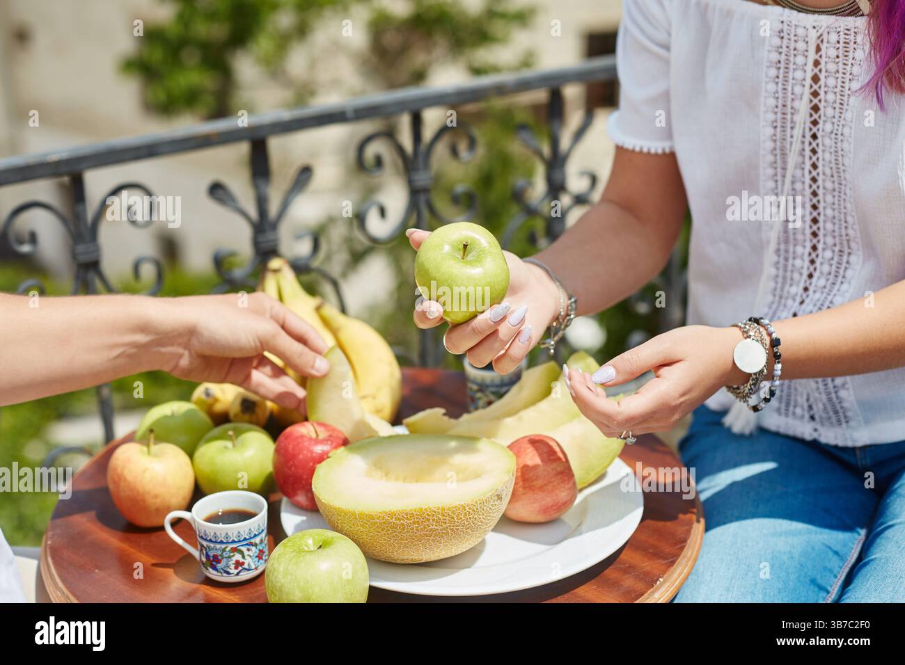 Due amici possono gustare una vivace selezione di frutta fresca su un balcone. Un amico tiene in mano una mela verde mentre è circondato da frutta colorata e una piccola tazza Foto Stock