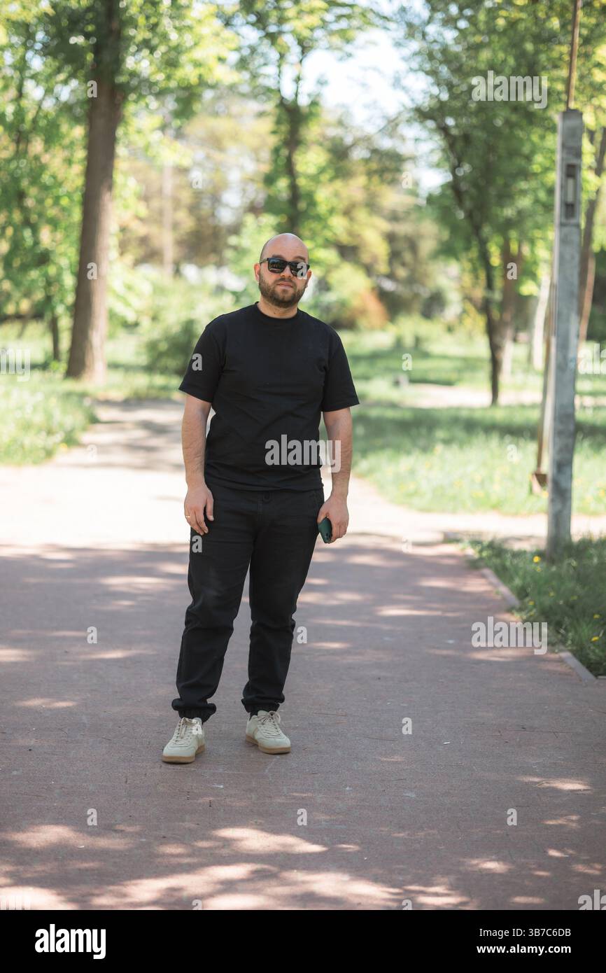 Ritratto in piedi di un uomo vestito con abiti casual con occhiali da sole. La foto viene scattata nel parco. Foto Stock