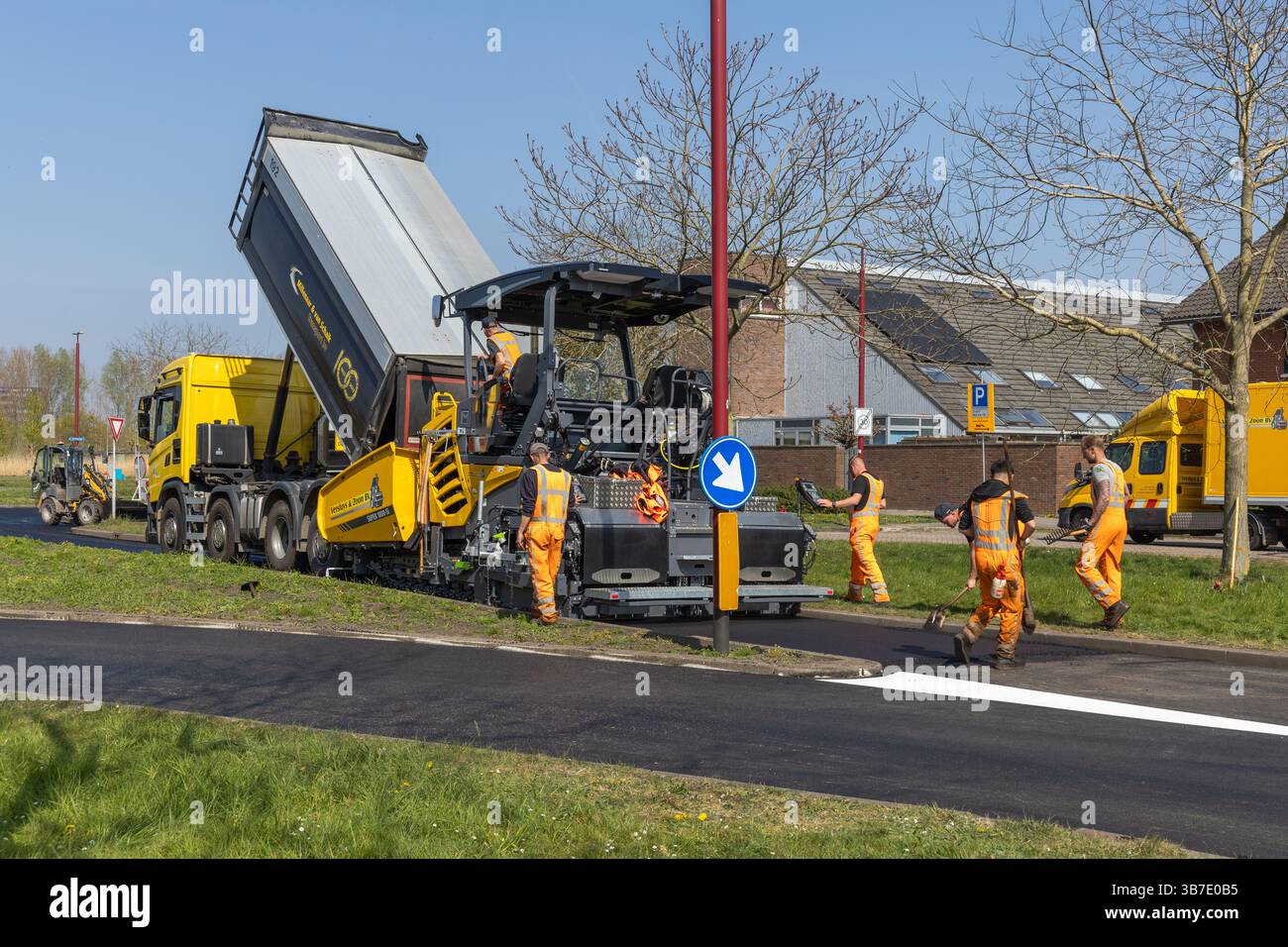 I lavoratori stradali in uniforme arancione gestiscono un'asfaltatrice e un camion durante una giornata di sole, aprendo nuovi marciapiedi in un'area urbana con lavoro di squadra e precisio Foto Stock