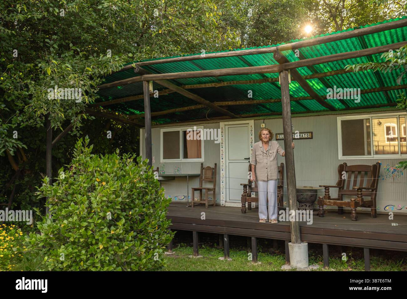 Donna anziana in pensione in una casa prefabbricata circondata da una natura lussureggiante, in piedi sul suo portico sotto un tetto di ferro ondulato verde Foto Stock