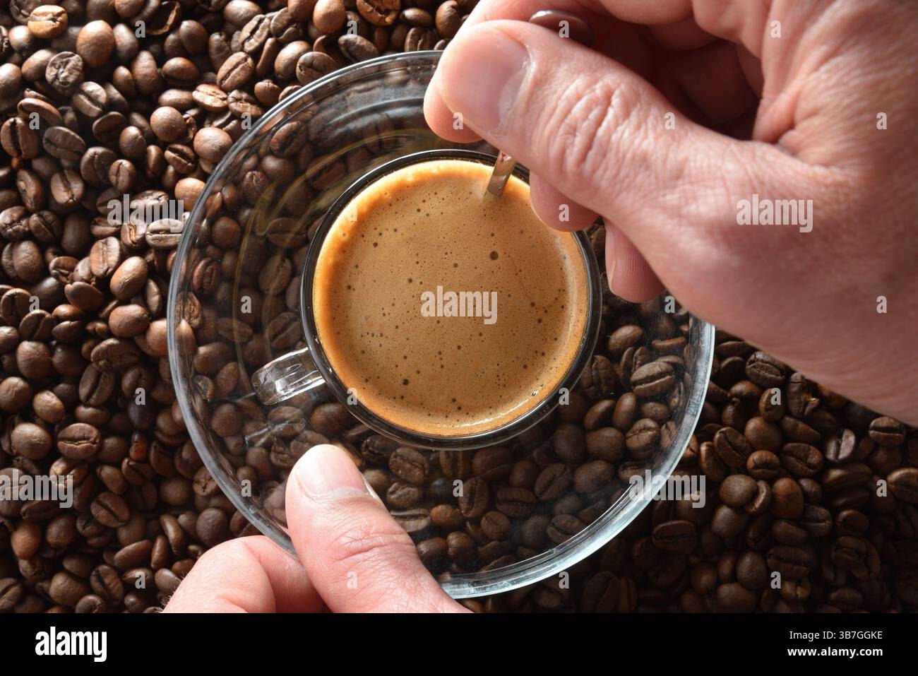 Dettaglio delle mani che mescolano il caffè appena estratto in una tazza di vetro e un piattino con un cucchiaio di metallo su una pila di chicchi di caffè. Vista dall'alto. Foto Stock
