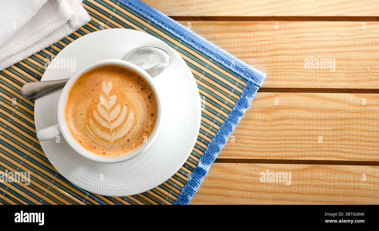 Caffè schiumoso con motivo floreale in tazza e piattino in ceramica bianca su una tovaglia di bambù su un tavolo di legno. Vista dall'alto. Foto Stock