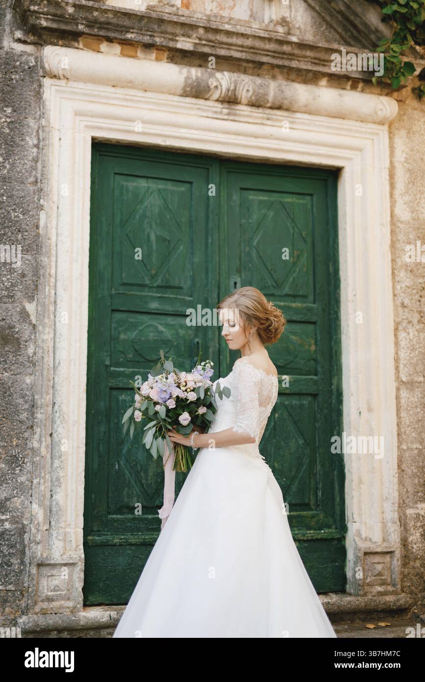 Una tenera sposa si erge con un bouquet in mano sullo sfondo di una porta verde di legno di un antico edificio. Foto Stock