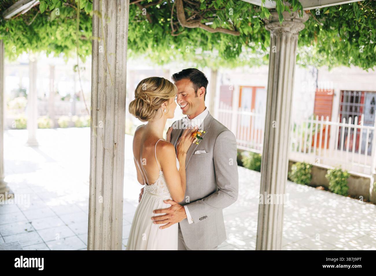 La sposa e lo sposo si stagliano tra le colonne, avvolgendosi e sorridendo. La sposa ha messo le mani sul petto dello sposo. . Foto Stock