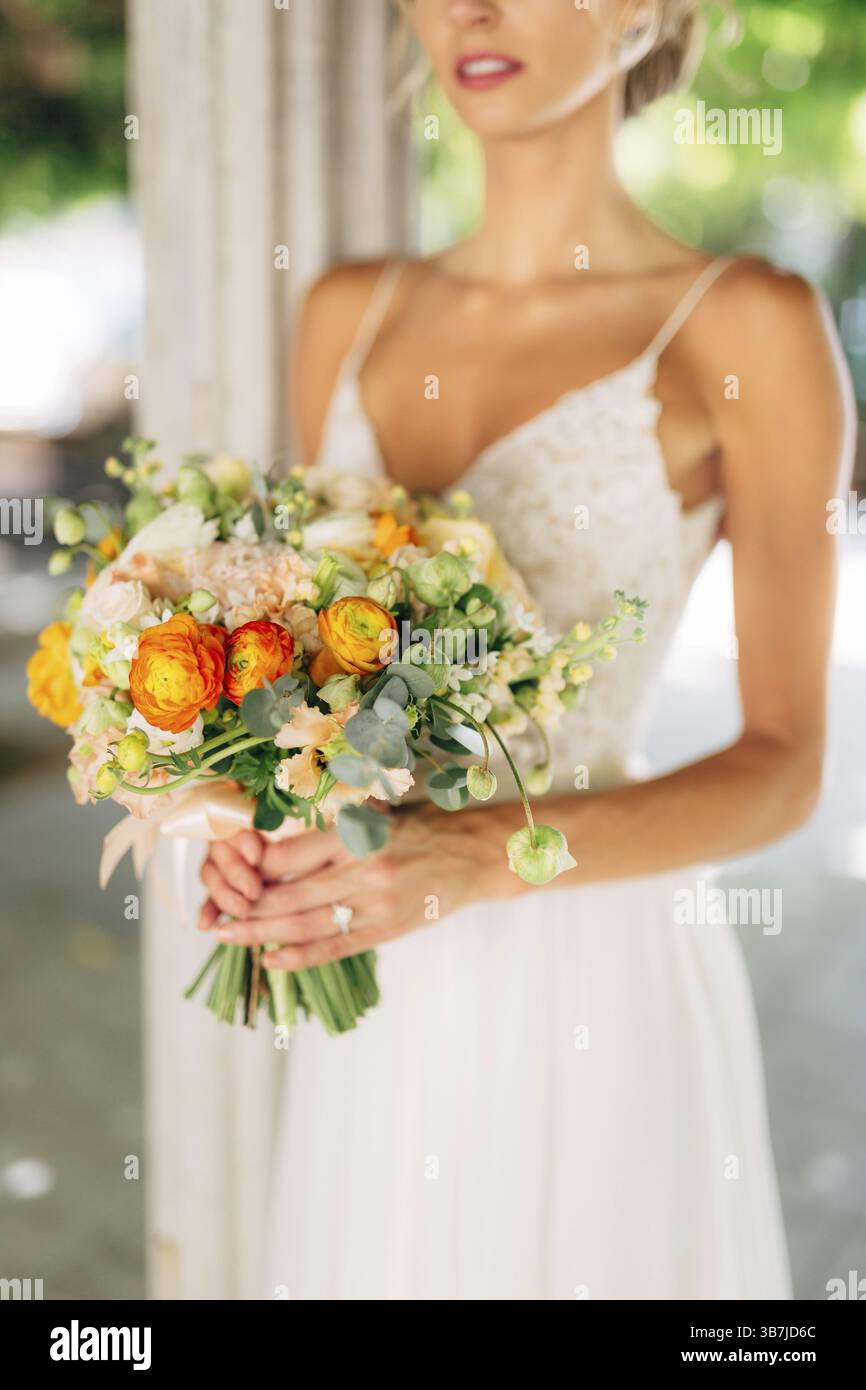 La sposa sorge tra colonne bianche intrecciate con viti verdi e tiene un bouquet nuziale con coppe arancioni, primo piano. Foto Stock