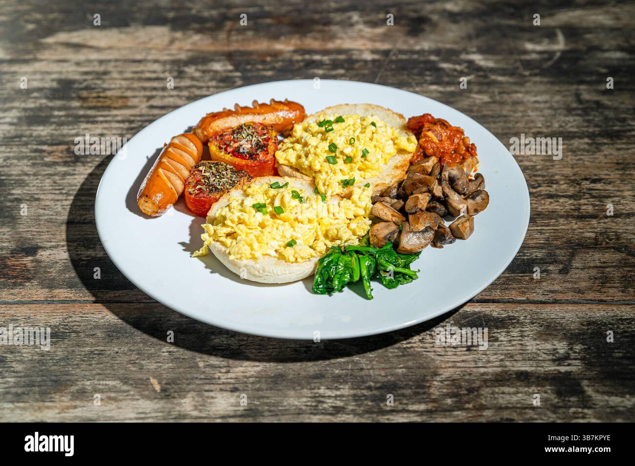 Colazione abbondante con pane, uova strapazzate, salsicce, pomodoro arrosto, fagioli al forno, funghi saltati e spinaci su piatto bianco. Isolato da un tavolo di legno Foto Stock