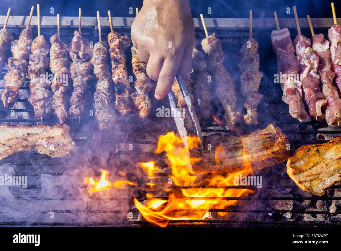 Una griglia ripiena di spiedini di carne che cucinano sopra le fiamme aperte. Il fumo aumenta man mano che la carne diventa marrone, mentre qualcuno tende alla griglia indossando i guanti. La sce Foto Stock