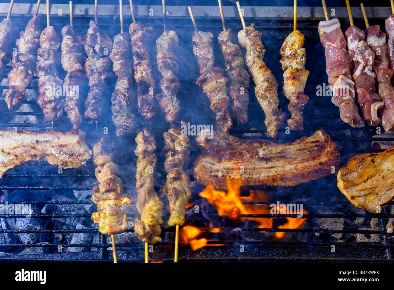 Una griglia ripiena di spiedini e pancetta di maiale di carne cucinata sopra le fiamme aperte. Il fumo aumenta man mano che la carne dorme, la scena mostra un barbecue tradizionale Foto Stock