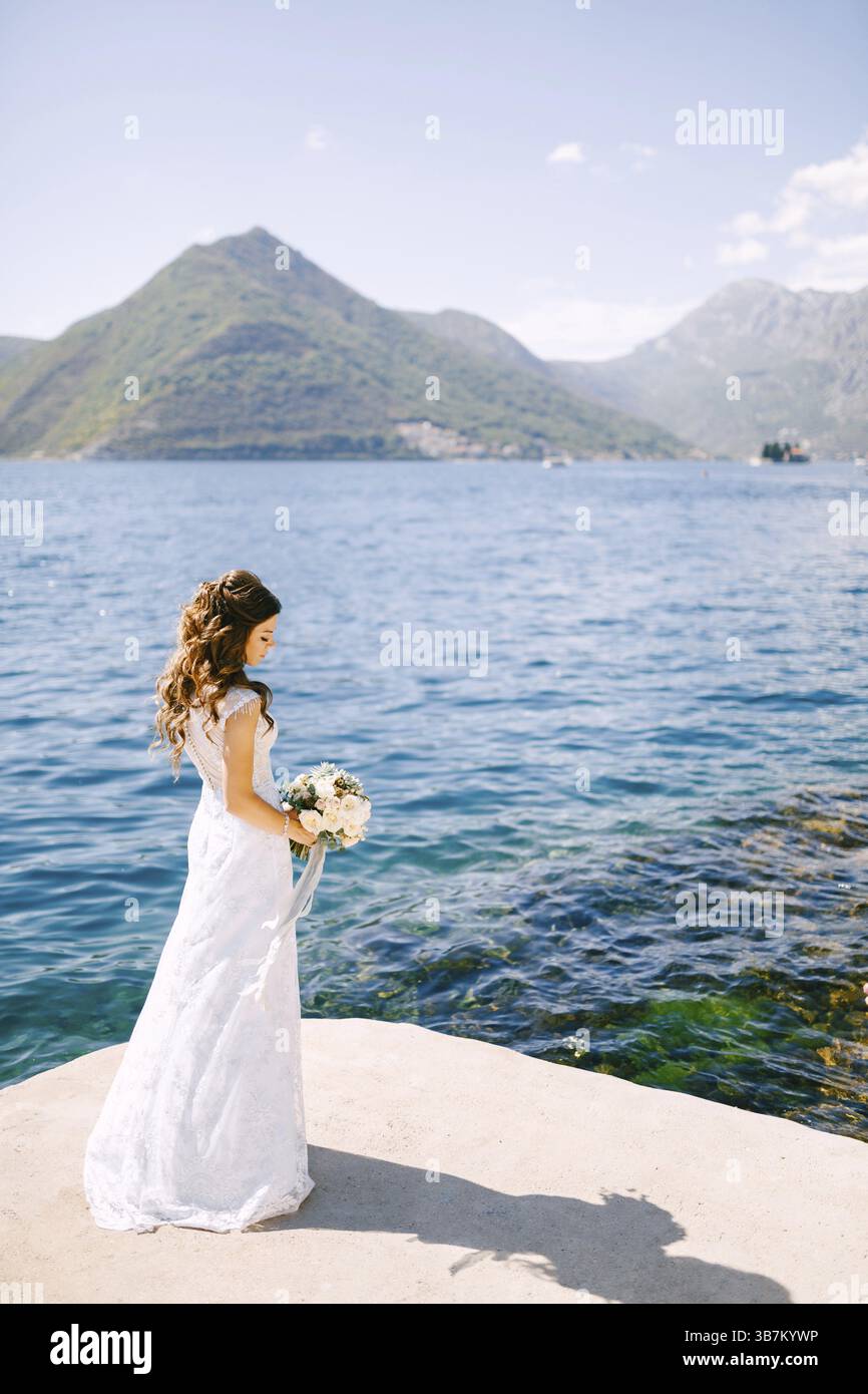 Una sposa elegante con un bouquet in mano si erge su un molo nella Baia di Cattaro vicino all'acqua blu trasparente. Foto Stock