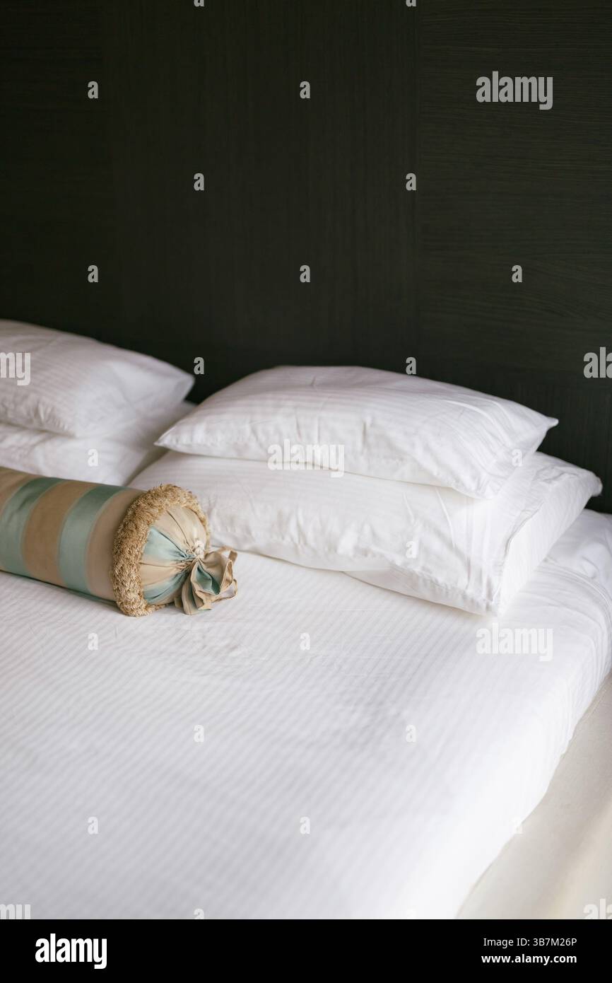Un letto con una coperta bianca e cuscini di diverse dimensioni e un cuscino allungato in una camera con pareti scure. Foto Stock