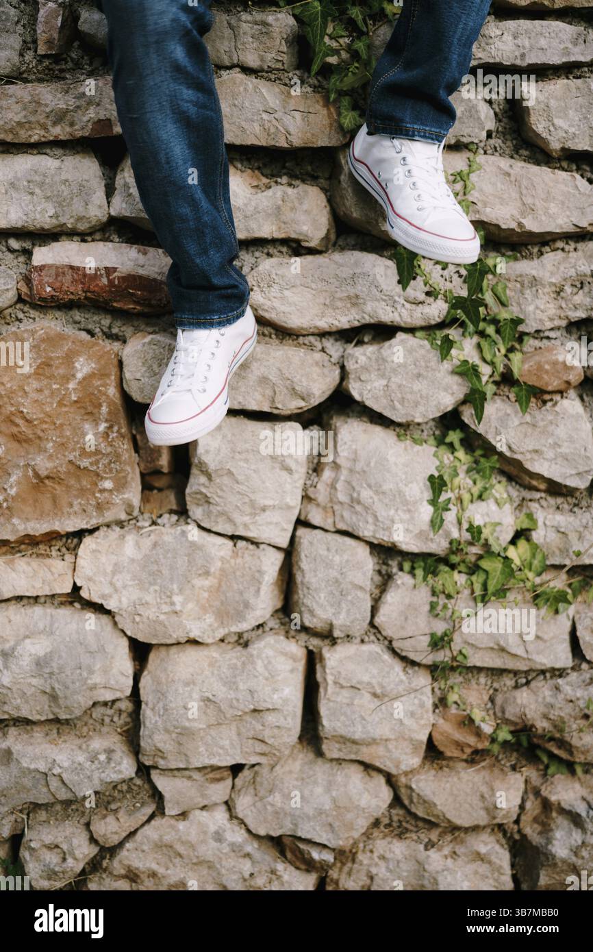 Gambe maschili in jeans e sneakers sullo sfondo di un muro di pietra nel parco. Foto Stock