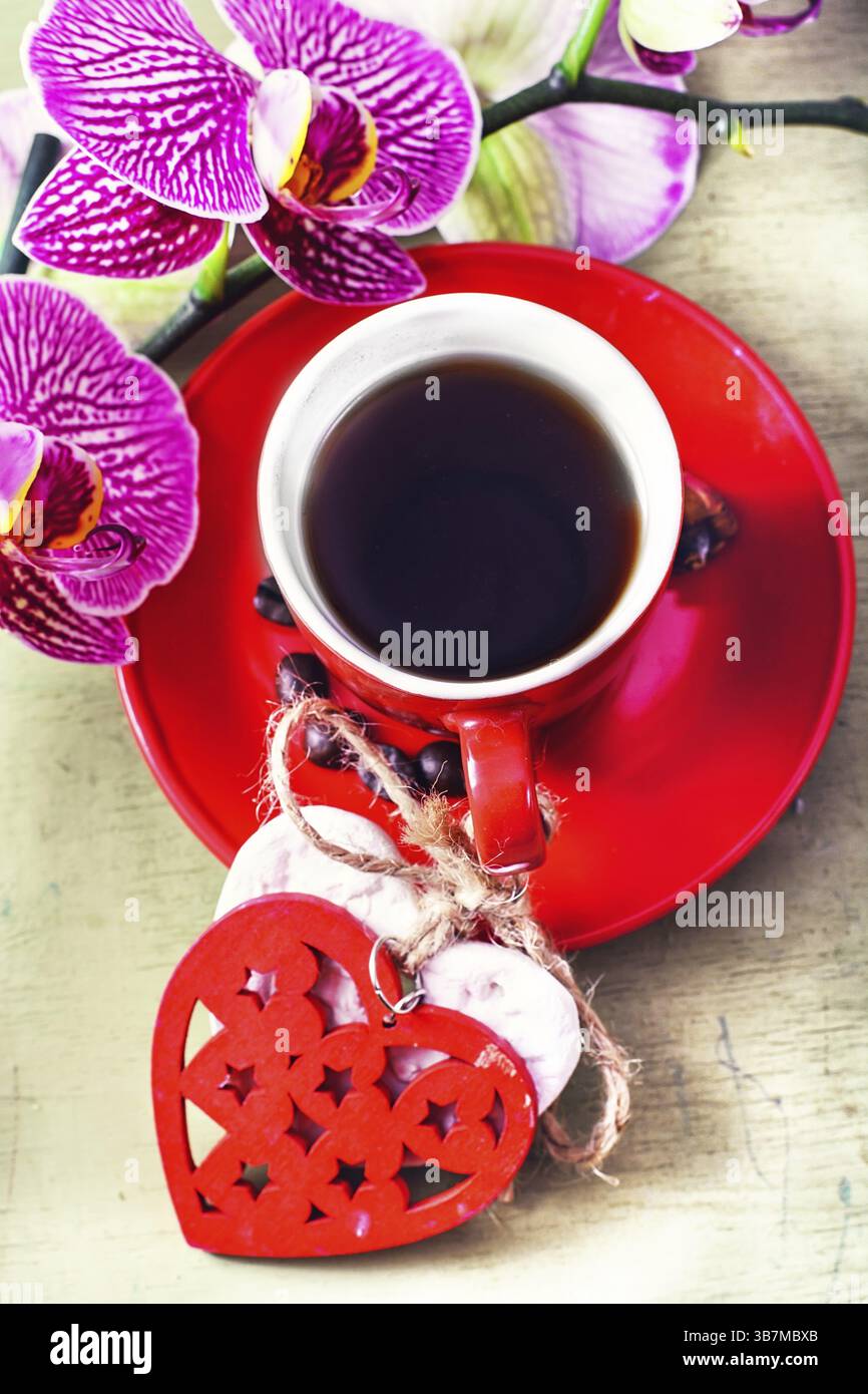 Tazza rossa decorata con cuori simbolici con caffè nero e un ramo fiorito di orchidee Foto Stock