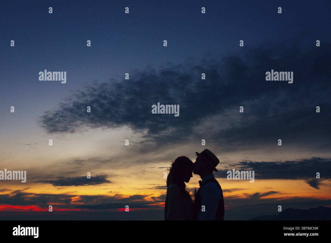 La silhouette dello sposo nel cappello bacia la sposa sulla fronte al tramonto. Foto Stock