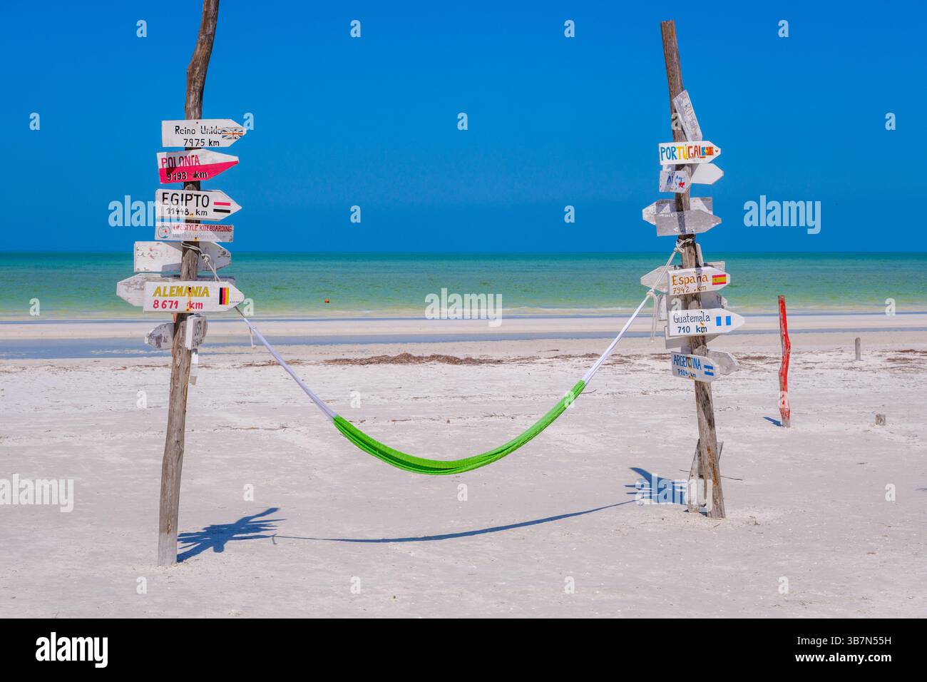 Holbox, Messico – 10 febbraio 2025: Un'amaca verde è legata tra due pali di legno ricoperti da cartelli direzionali che puntano verso paesi lontani, set Foto Stock
