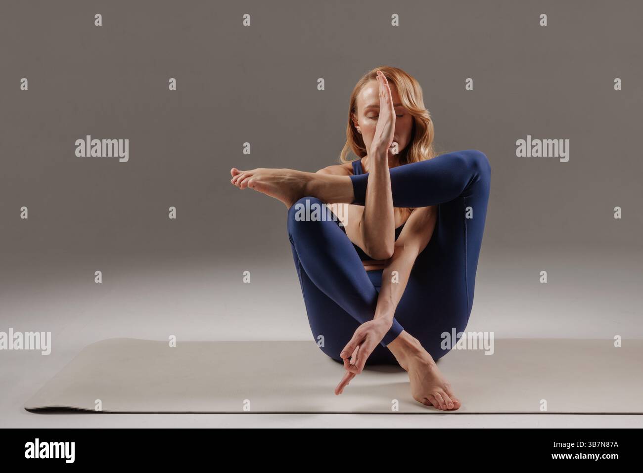 Donna flessibile che esegue una posa yoga complessa con una concentrazione profonda, dimostrando un controllo avanzato del corpo in ambienti interni. Foto Stock