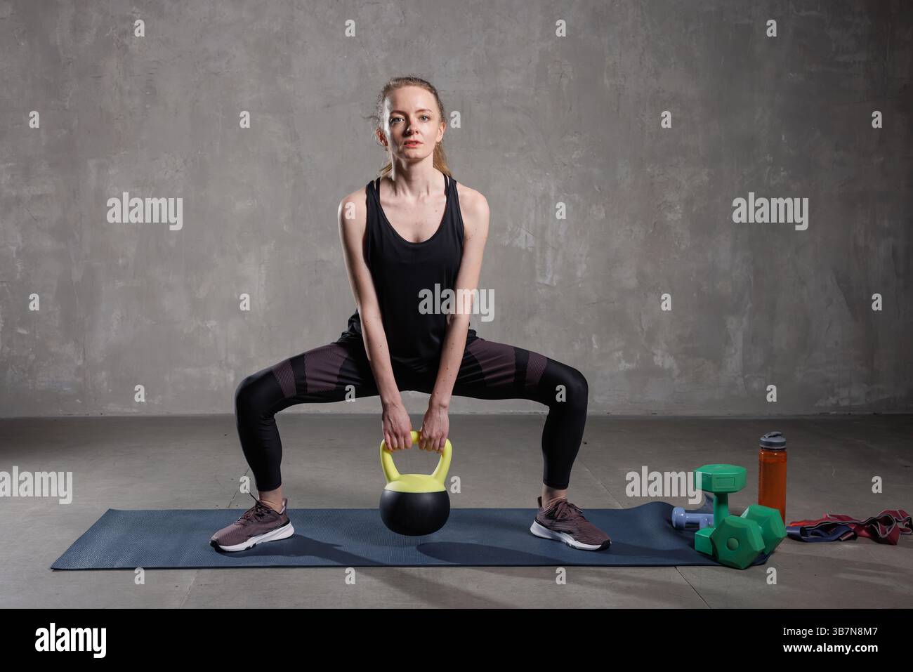 Fit Woman che esegue uno squat di sumo kettlebell su un tappetino da yoga in palestra, concentrandosi su forza, equilibrio e tonificazione del corpo. Foto Stock