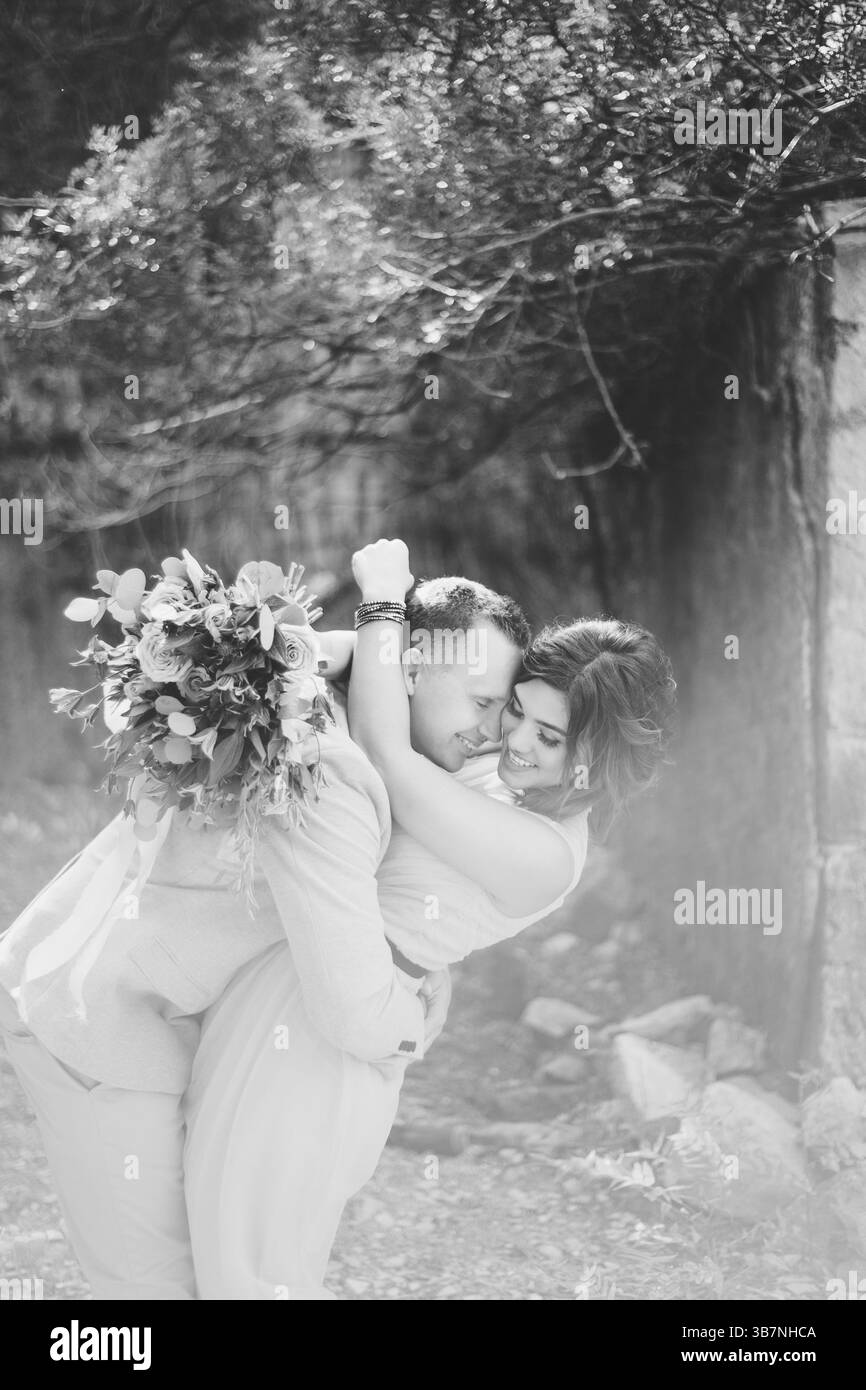 Foto in bianco e nero. Lo sposo felice sorridente abbraccia la sposa in un bel vestito. La sposa tiene tra le mani un grande bouquet di fiori. Foto Stock