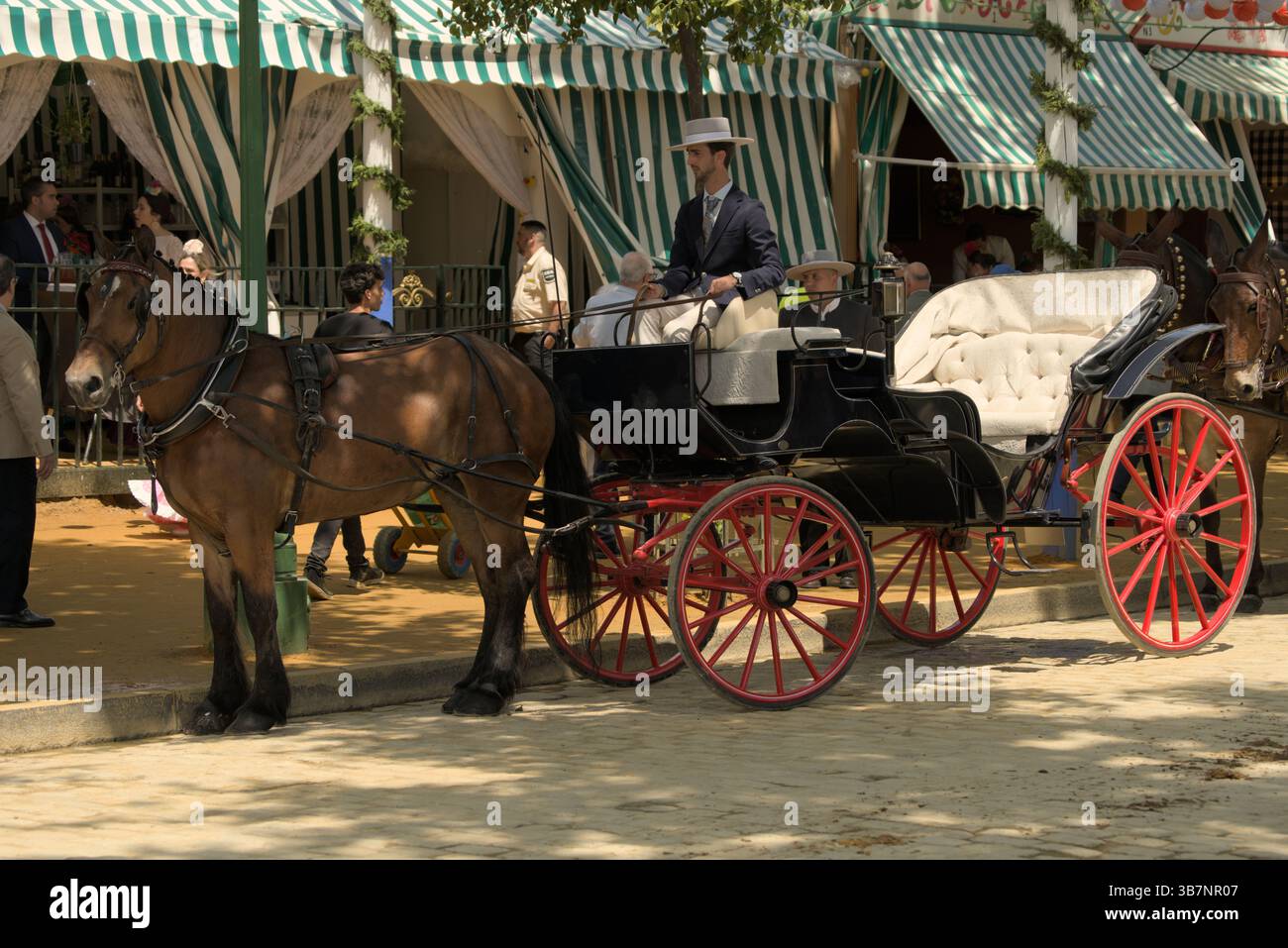 Elegante carrozza andalusa con ruote rosse a la Feria de Sevilla 2025, di fronte alle casetas a righe.Feria de Sevilla, Spagna Foto Stock