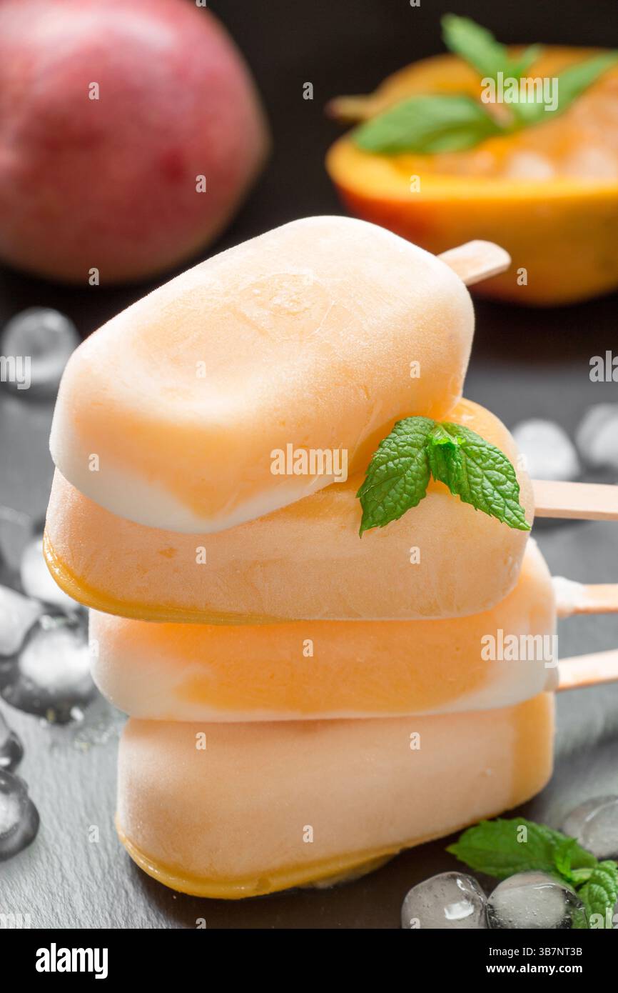 Colonna di 4 sundae di sorbetto di mango mescola il gelato dietro il mango e il mango tagliato vicino ai cubetti di ghiaccio su sfondo nero. Mango sorbetto sundae mescolare i papaveri del gelato. Verticale. Foto Stock