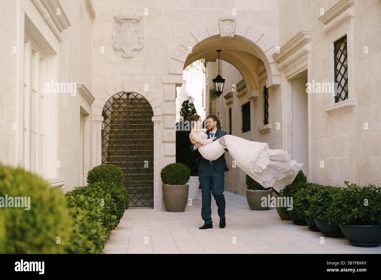 Lo sposo porta la sposa tra le braccia nel cortile della casa con archi. Foto Stock