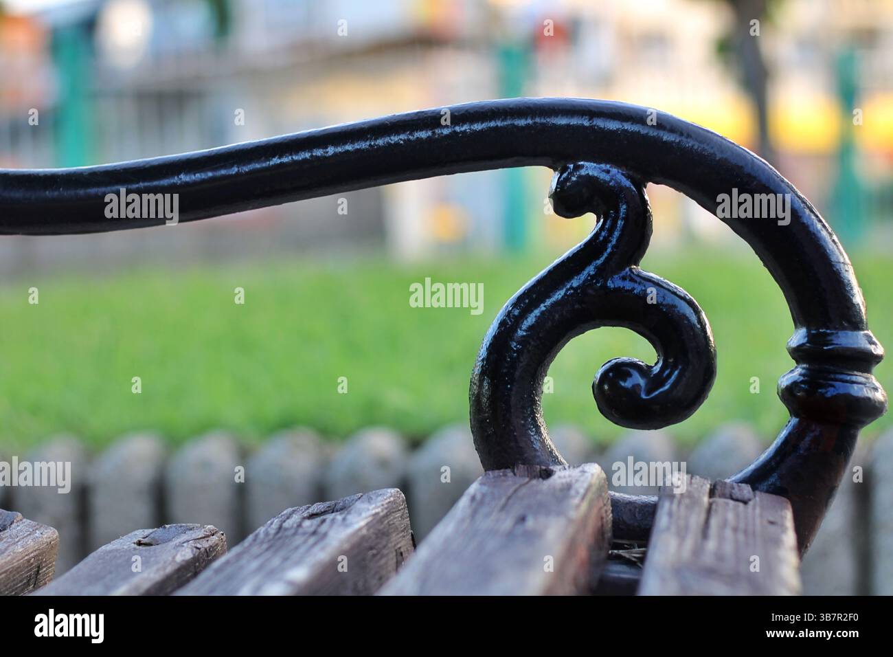 Primo piano di braccioli decorativi in ferro battuto e spranghe in legno invecchiato su una panchina da parco d'epoca, con erba sfocata e sfondo del parco. Foto Stock