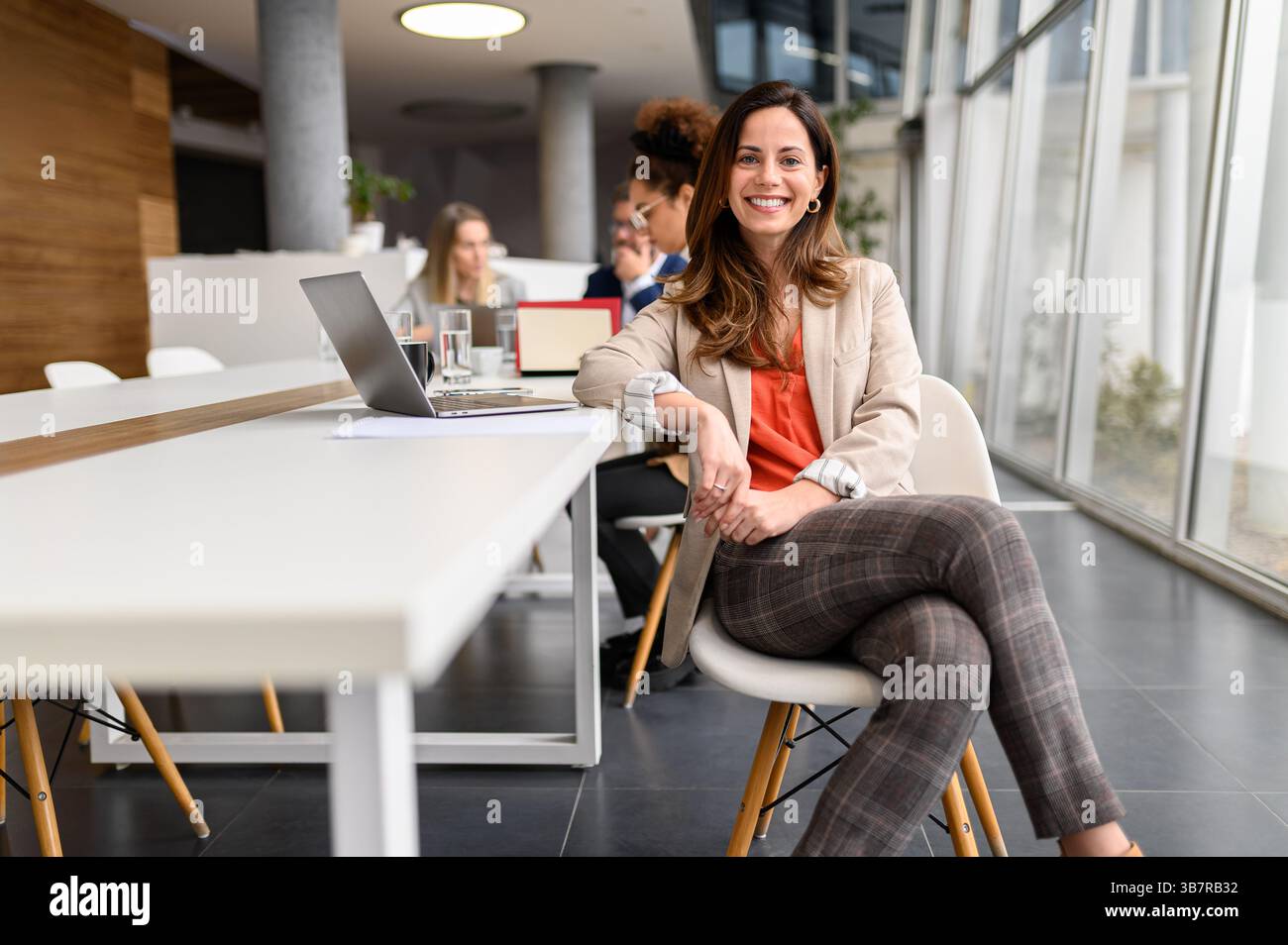 Ritratto di una donna d'affari sicura di sé con un computer portatile al tavolo seduto nella sala riunioni mentre i colleghi collaborano sullo sfondo Foto Stock